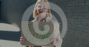 Slow motion of mature woman activist holding It's our future sign and smiling standing against brick wall background