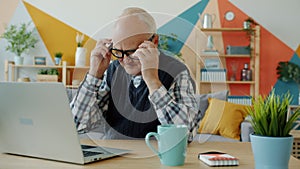 Slow motion of old man putting on eyeglasses then working with laptop in apartment