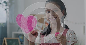 Slow motion portrait of adorable child smiling painting pink heart on glass board looking at camera