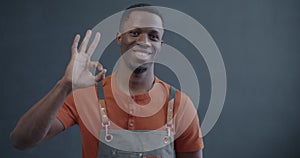 Slow motion portrait of African American man wearing apron showing OK hand gesture and smiling on gray background