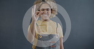 Slow motion portrait of African American woman in apron entrepreneur showing OK hand gesture and winking on grey