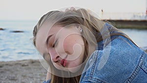 Slow motion portrait of cheerful girl who poses on camera and cute laughs sitting on sand on background of blue sea and