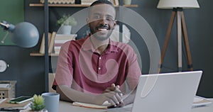 Slow motion portrait of cheerful young African American man employee sitting at desk in office smiling