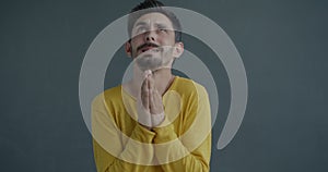 Slow motion portrait of emotional young man praying begging putting hands together looking up on grey background