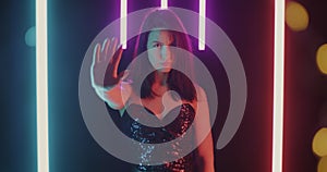 Slow motion portrait of gloomy young woman making Stop gesture stretching arm to camera on glowing neon background