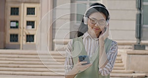 Slow motion portrait of happy Asian woman listening to music with headphones holding smartphone walking in city