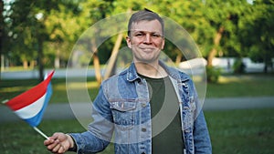 Slow motion portrait of joyful guy happy tourist waving flag of the Netherlands, smiling and looking at camera. National