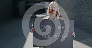 Slow motion portrait of mature woman holding You are not alone sign expressing solidarity standing against brick wall