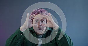 Slow motion portrait of shocked young man staring at camera through imaginary binoculars on purple background