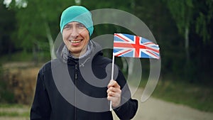Slow motion portrait of smiling Englishman sports fan holding UK flag flying in the wind and looking at camera