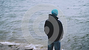 Slow motion portrait of young man tourist standing on the sea shore and looking at waves on cold windy day. People