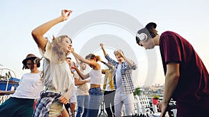 Slow motion of pretty young woman in sunglasses dancing on rooftop with her friends when deejay in wireless headphones