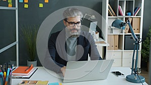 Slow motion of serious mature man putting on glasses then working with laptop in office