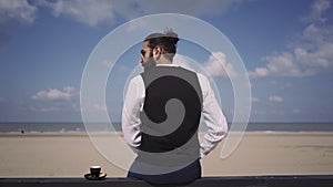 Slow-motion shot of male back profile on the beach manbun beard eyeglasses coffee cup plate sitting