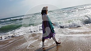 Slow motion side view of Caucasian blonde woman in pink dress walking on sandy beach, sea waves washing the shore, summer vacation