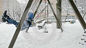 Slow motion of smiling boy swings on a playground swing covered in snow at a winter park. Concept of childhood.