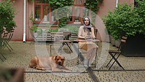 Slow motion. Stylish woman smiling and drinking morning coffee while sitting at a table in a city outdoor cafe on a