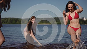 Slow motion. Three bikini girls run into the river and cheerfully splash water on each other.