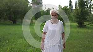 Slow motion. A tired elderly upset woman in a white dress leisurely walks along the green grass in a city park on a