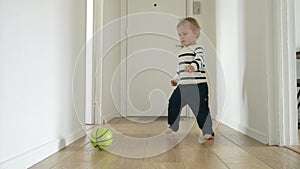 Slow motion of a toddler boy as he plays with a ball on the wooden floor of a long corridor in his house