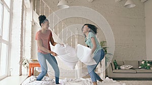 Slow motion of two mixed race young pretty girls jumping on bed and fight pillows having fun at home