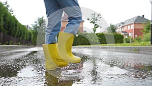 Slow motion video of little boy wearing yellow rubber boots jumping in puddle of water on rainy summer day in small town.