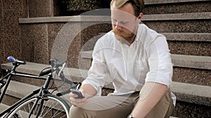 Slow motion video of stylish young man sitting on stone staircase and using mobile phone