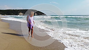 Slow motion view of gorgeous Caucasian blonde woman in pink dress walking on sandy beach, sea waves washing the shore