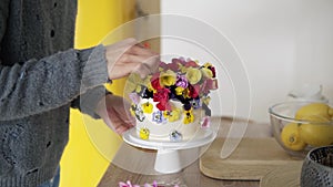 Slow motion of woman with short grey hair decorates small white cake with colorful flowers on white modern kitchen