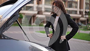 Slow motion. A woman stands in front of the open hood of a car and looks at the engine in confusion, taking a deep
