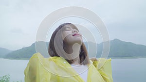 Slow motion - Young Asian woman feeling happy playing rain while wearing raincoat standing near lake.