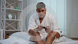 Slow motion. A young guy in a white bathrobe with a joystick in his hands plays on a console while sitting on a bed in