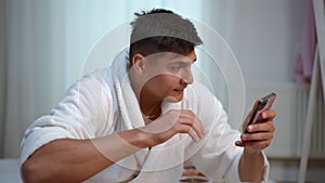 Slow motion. A young guy in a white bathrobe sits on a bed in the bedroom, drinks water from a glass and looks at cell