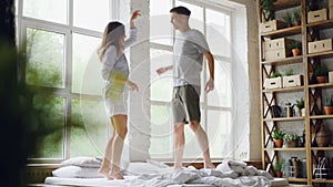 Slow motion of young married couple jumping and dancing on double bed in light room with large windows, happy people are