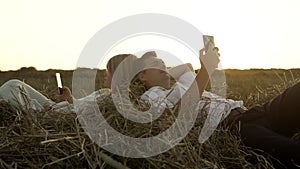 Slowmo of caucasian couple who is lying on haystack in field at sunset and watching their mobiles