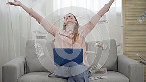 Slowmo of confident young woman pressing key on laptop keyboard and money falling on her from above. Slow motion