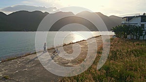 Slowmotion aerial shot. Woman and her son ride kick scooterand bicycle by the sea during sunset