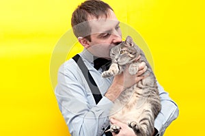 Sly man in blue shirt holding cute grey tabby cat and jesting bite it ear in front of yellow background