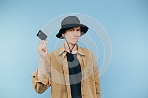 Sly young hipster man in panama and shirt stands on a blue background and shows a credit card to the camera with a smile on his