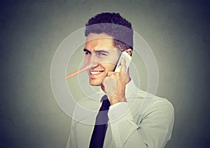 Sly young man with long nose talking on mobile phone on gray wall background. Liar concept.