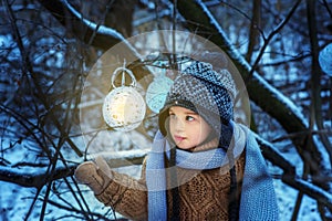 Small 4 yaers old girl in snowy winter forest looks on luminous ball. Child in brown pullover, gray scarf and warm hat. Christmas