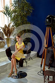 A small 6-year-old girl sits on a chair, pampas grass, palm tree, designer furniture.Yellow dress in peas and a bun