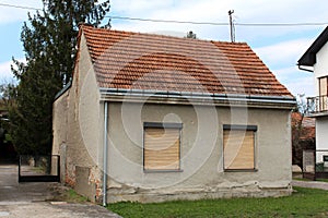Small abandoned family house with dilapidated facade and two front windows covered with window blinds surrounded with grass and