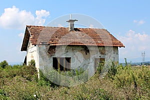 Small abandoned red bricks house with dilapidated white facade and destroyed roof tiles completely surrounded with high grass