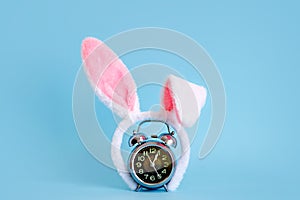 Small alarm clock and bunny ears on pastel blue background. Eastertime minimal concept