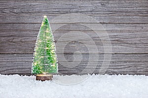 small artificial green christmas tree with holiday lights on snow against wooden rustic background