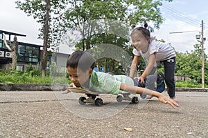 Small asain teenage girl playing with brother pushing forward boy riding on skateboard in public road. Kids and entertainment.
