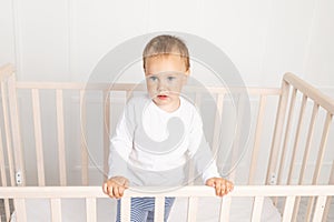 Small baby boy 2 years old in the crib looking at the camera in the bright children`s room, the child after sleeping