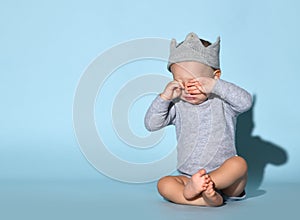 Small baby boy in jumpsuit, decorative crown on head and barefoot sitting on floor, touching eyes and crying or feeling sleepy