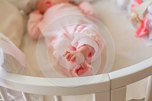 Small baby feet clad in pink romper. Tiny fingers of the newborn. The child has crossed legs and lies in a round white bed